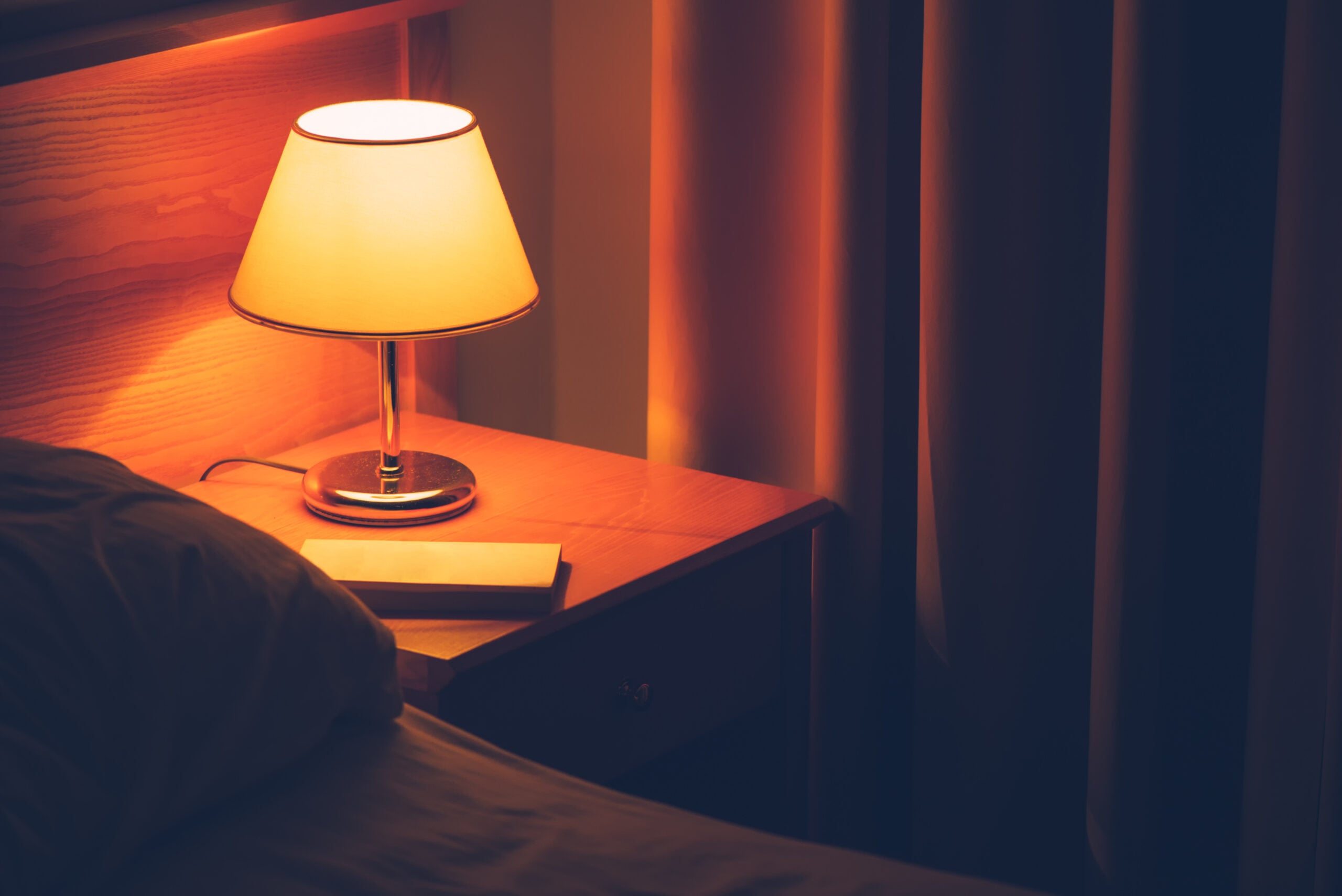 A warm bedside lamp glowing next to a book on a wooden nightstand at dusk