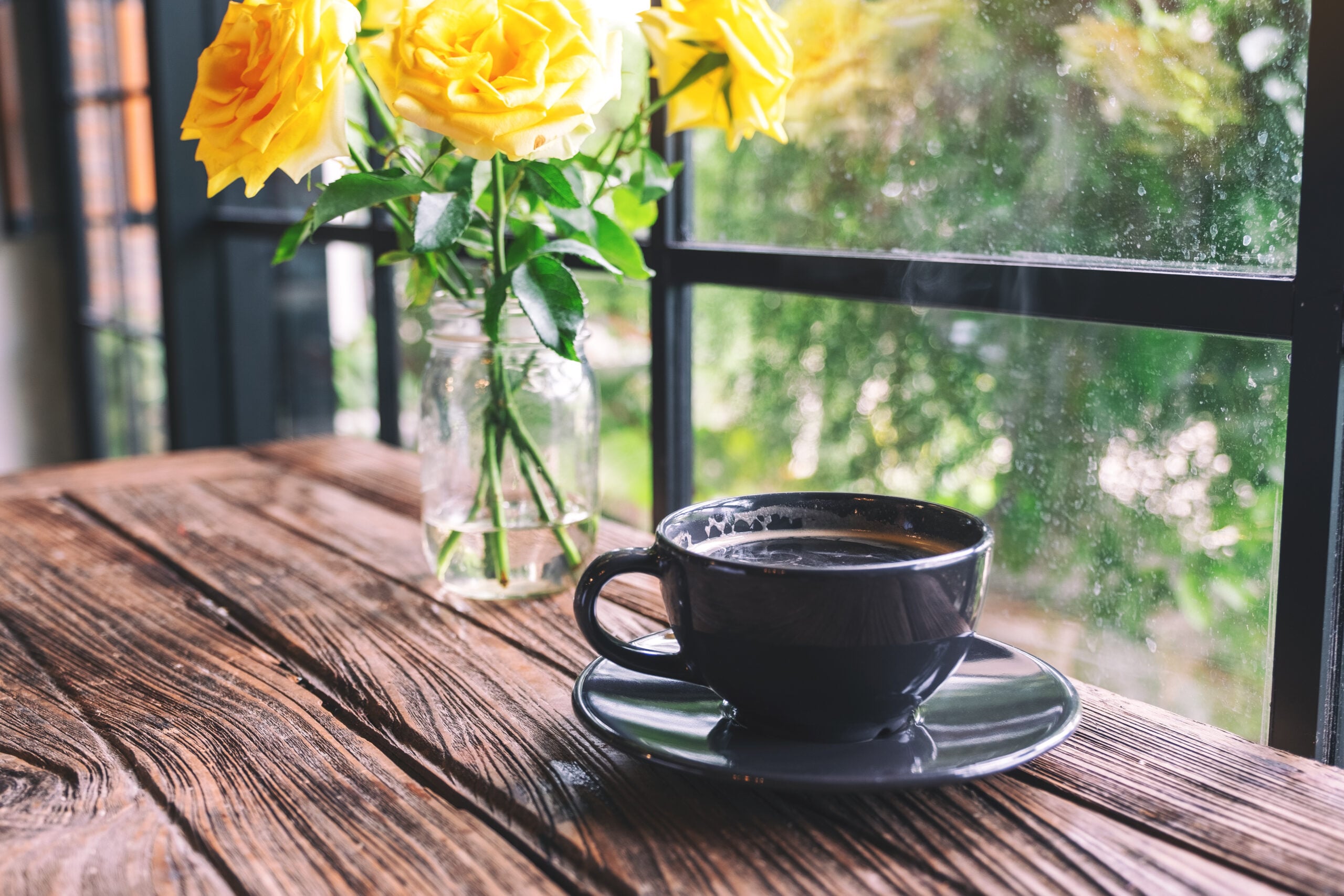 A cup of hot black coffee and yellow roses on a vintage wooden farmhouse table in warm morning light