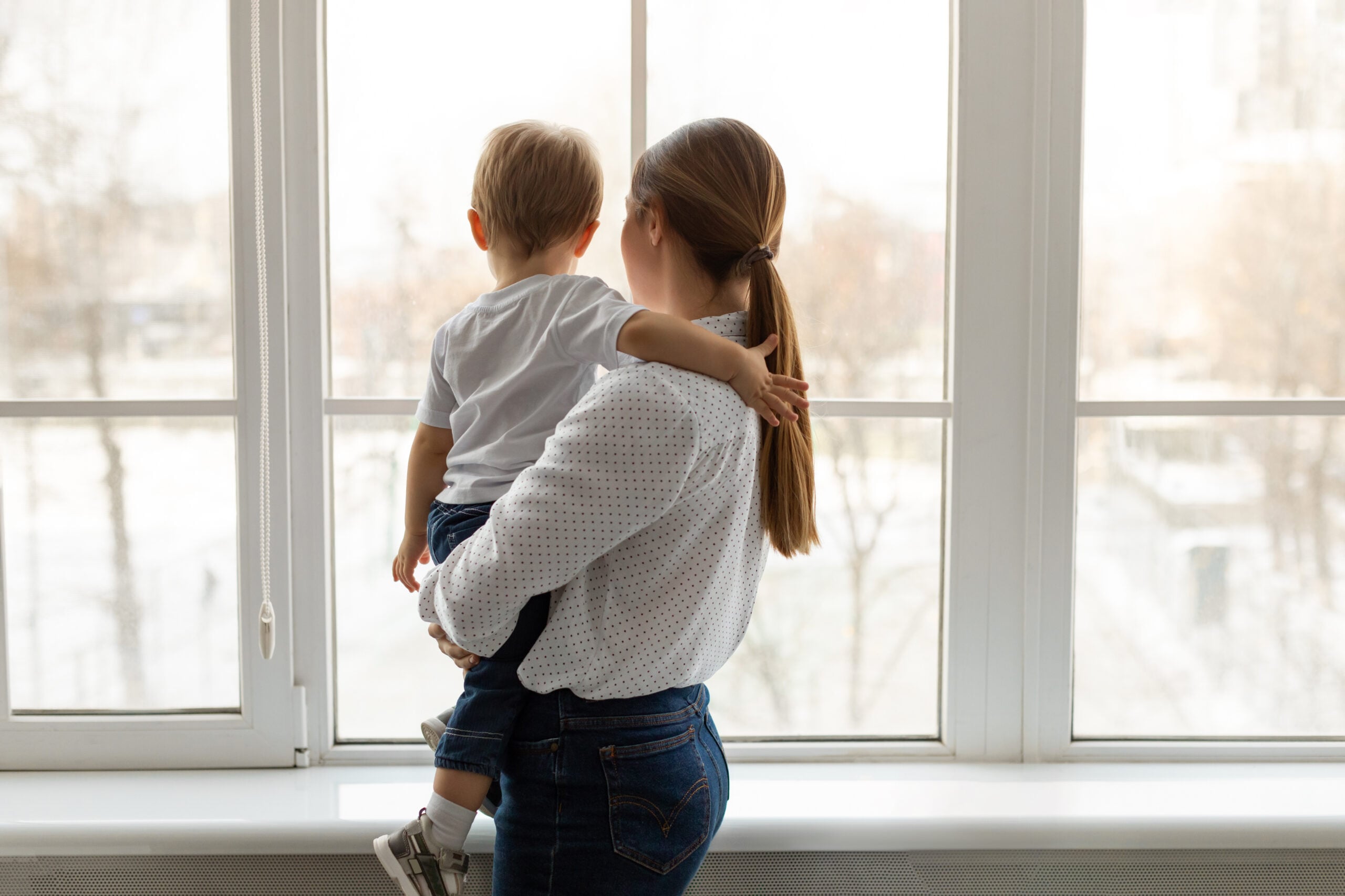 A parent holding a toddler at a window, seen from behind, looking out at the yard