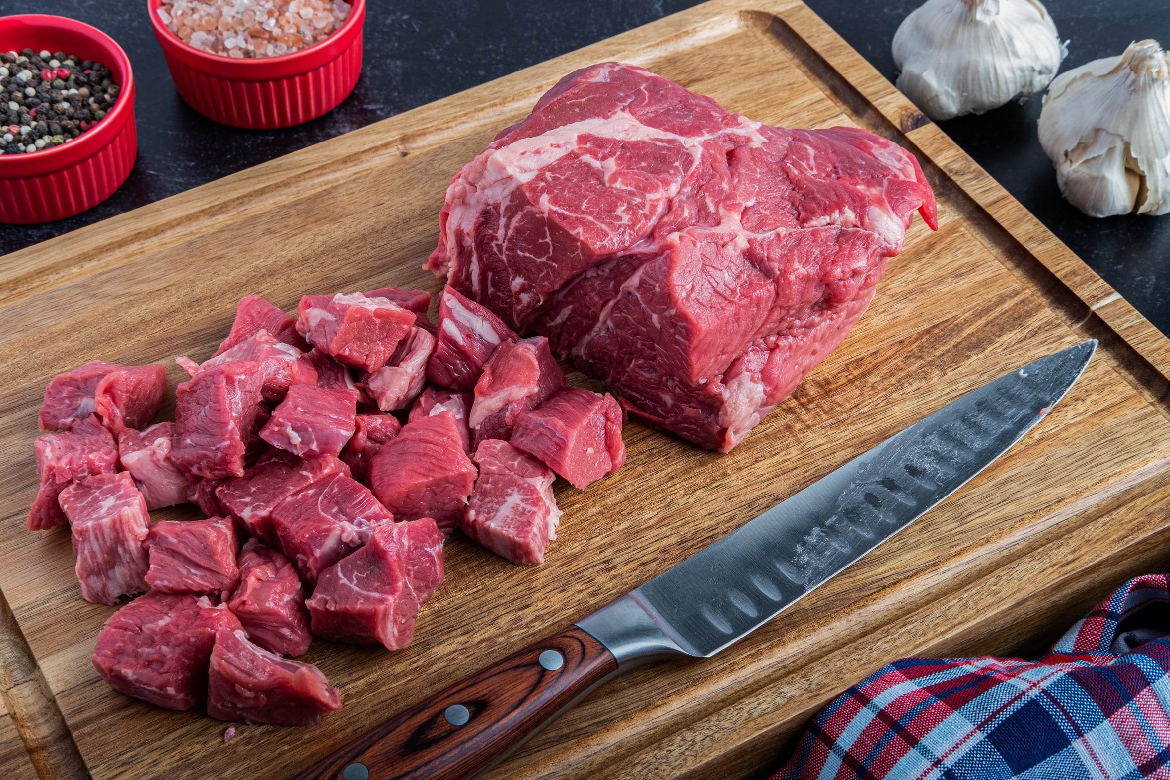Boneless beef chuck roast cut into cubes on a wooden cutting board with salt and peppercorns