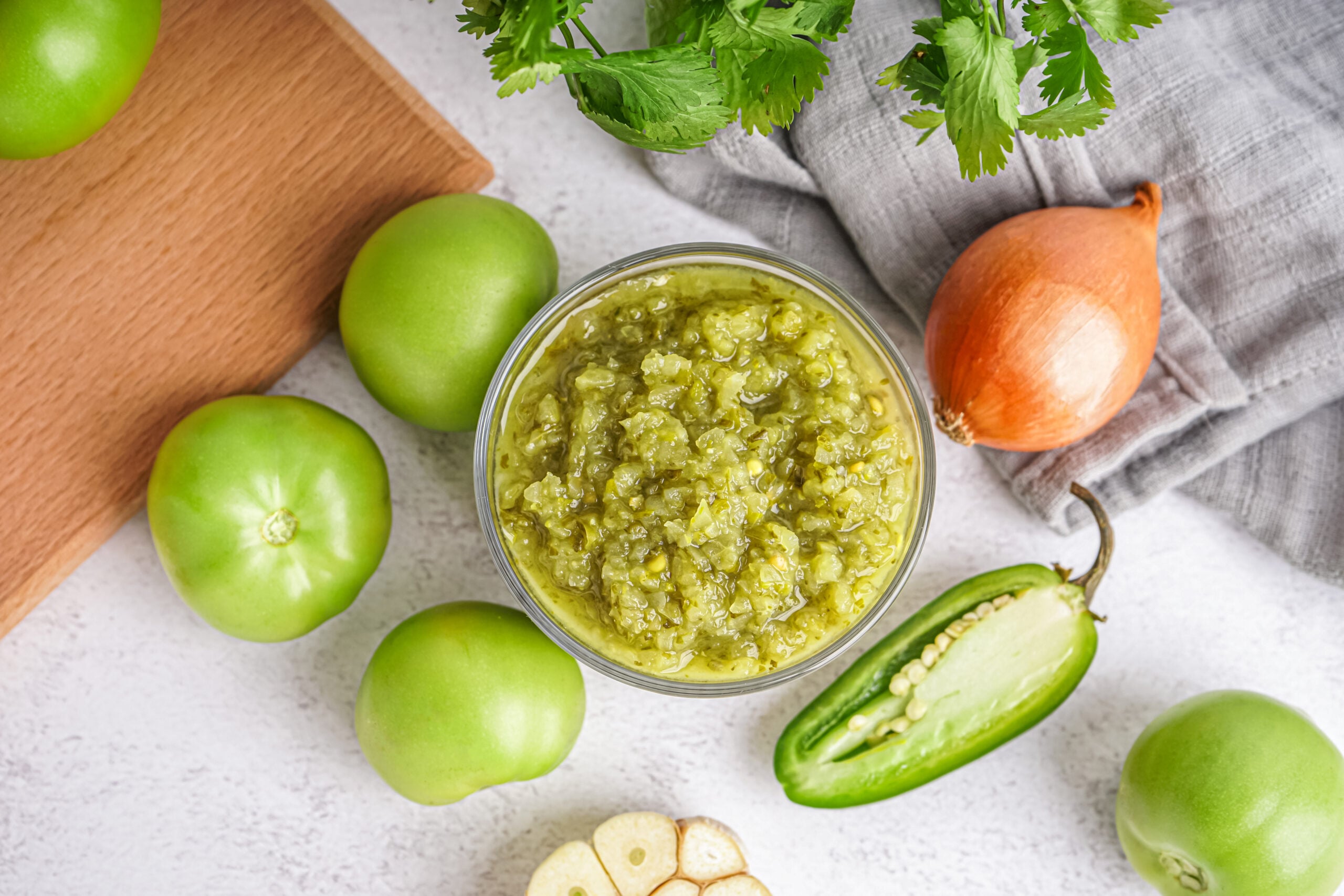 A bowl of roasted green tomato salsa surrounded by whole green tomatoes, onion, jalapeno, garlic and cilantro